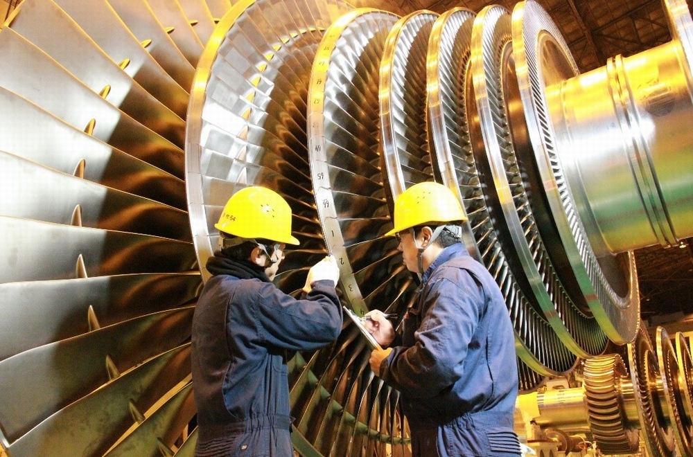 Kỹ sư đội mũ bảo hộ đang kiểm tra một cụm stato/máy phát cỡ lớn trong nhà xưởng, với máy tính giám sát đặt bên cạnh